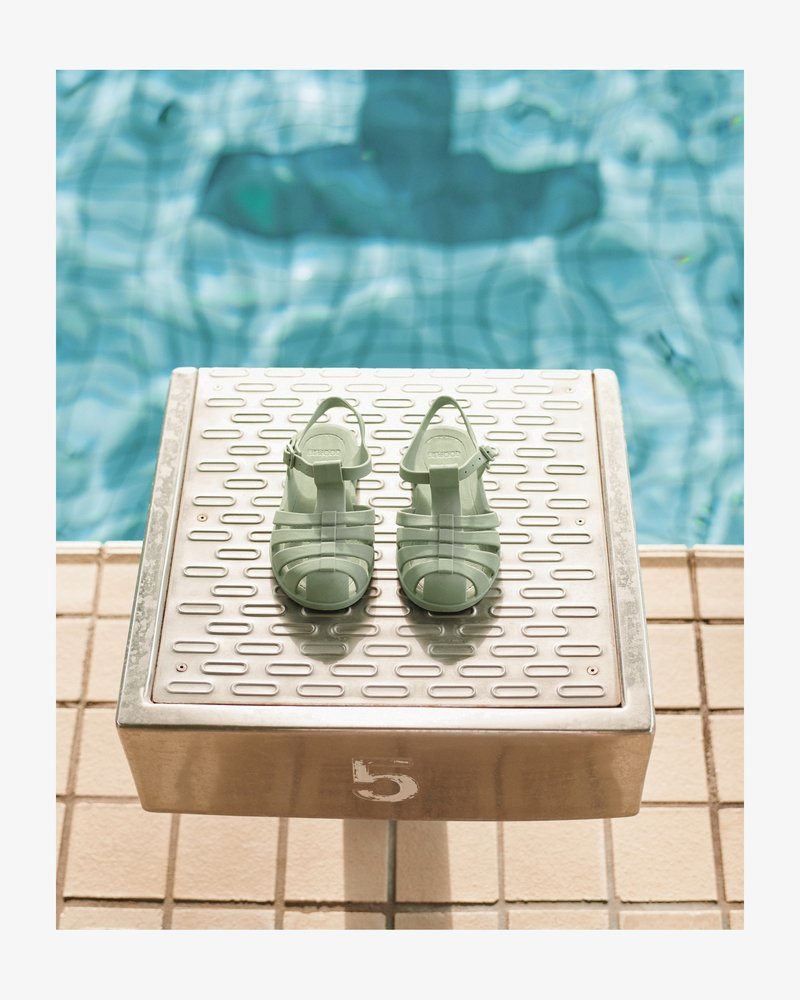 Sandales vert menthe translucides avec un design à lanières et un bout ouvert, posées sur une plateforme métallique près d'une piscine.