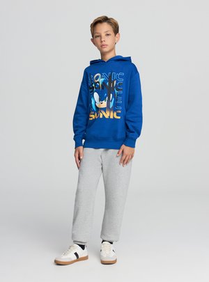 Felpa con cappuccio blu con grafica di Sonic, pantaloni della tuta grigi e sneakers bianche con dettagli neri e marroni. Tessuto morbido, vestibilità comoda.