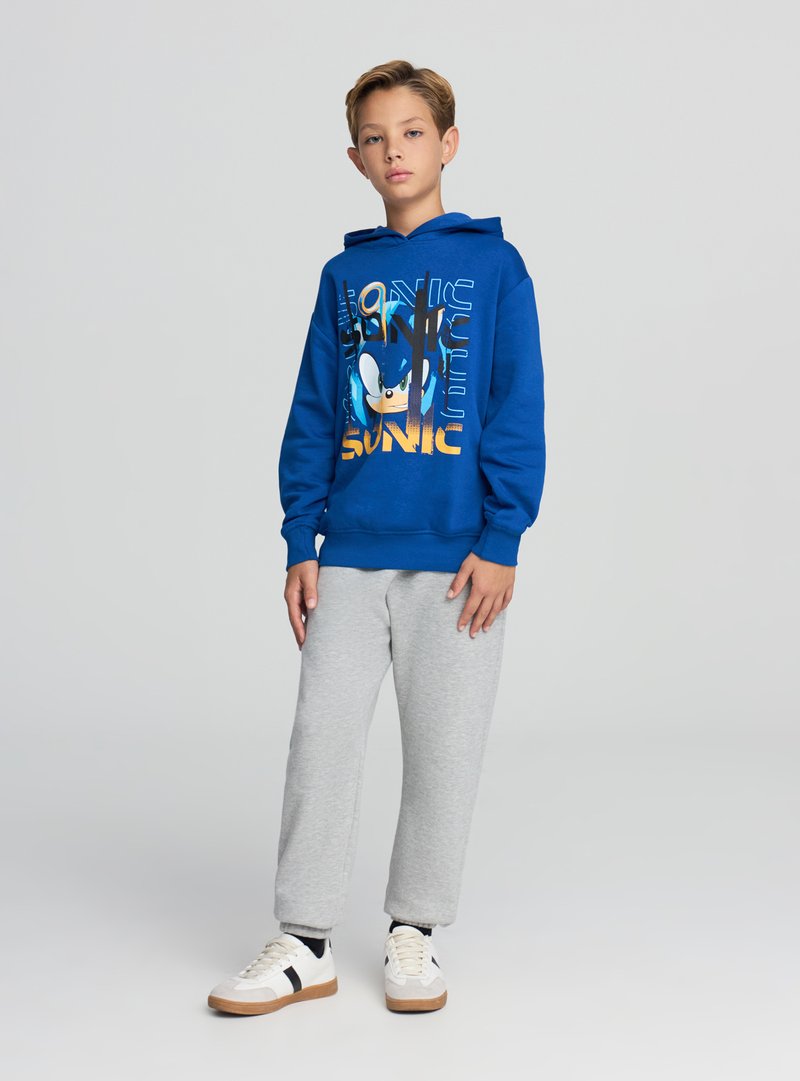 Felpa con cappuccio blu con grafica di Sonic, pantaloni della tuta grigi e sneakers bianche con dettagli neri e marroni. Tessuto morbido, vestibilità comoda.