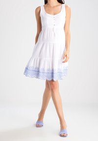 Vestido blanco sin mangas con un diseño en capas, que presenta adornos bordados en azul en el dobladillo. Botones en la parte delantera, hecho de una tela ligera.