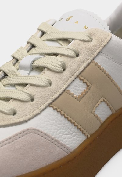 Gros plan d'une basket blanche et beige avec du cuir texturé, des accents en daim, des lacets beige et une semelle en caoutchouc gomme.