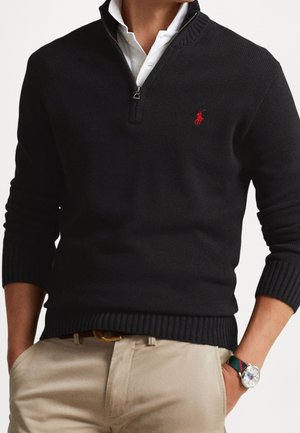 Mann trägt schwarzen Viertel-Reißverschluss-Pullover über weißem Hemd mit Kragen, beige Hose, brauner Gürtel und Armbanduhr mit gestreiftem Stoffarmband.