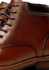 Brązowy skórzany but o gładkiej fakturze, z złotymi oczkami, ciemnymi sznurowadłami oraz wzmocnionym fasonem wokół kostki z kontrastującymi brązowymi akcentami.