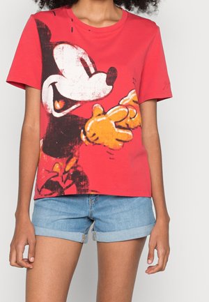 Henkilö, jolla on päällään punainen T-paita, jossa suuri retrohenkinen Mikki Hiiri -kuvio, ja vaaleansiniset farkkushortsit, joiden lahkeet on kääritty ylös.