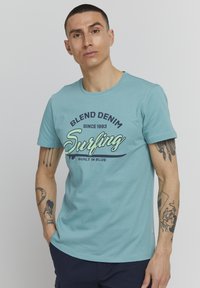 Gaišs teal kokvilnas T-krekls ar apaļu kakla izgriezumu, īsām piedurknēm un drukātu dizainu ar vārdiem "BLEND DENIM Surfing BUILT IN BLUE" kontrastējošās krāsās.