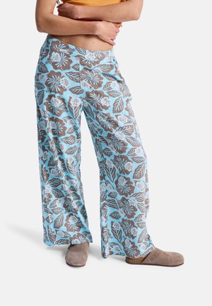 Persoon draagt losse blauwe broek met bruin-witte bloemenprint, beige slip-onschoenen en een cropped oranje top met gekruiste armen.