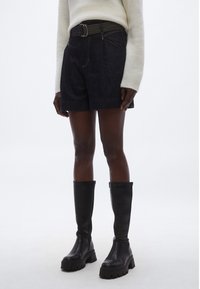 Črne kratke hlače iz visokokvalitetnega jeansa z visokim pasom in rumenim šivom, opremljene s pasom, v kombinaciji z visokimi črnimi usnjenimi škornji z debelimi podplati.