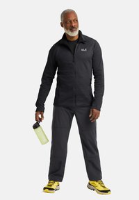 Giacca nera con zip, pantaloni grigio scuro e scarpe da escursionismo gialle brillanti. Tiene in mano una bottiglia d'acqua verde. Abbigliamento casual per attività all'aperto.