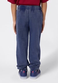 Kind in blauwe sweatpants en bijpassende blauwe sneakers, met de rug naar de camera gericht tegen een effen achtergrond.