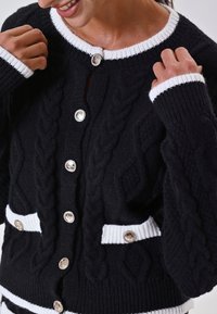 Cardigan nero lavorato a maglia con accenti a righe bianche, caratterizzato da motivi a treccia, bottoni argento e due tasche frontali.