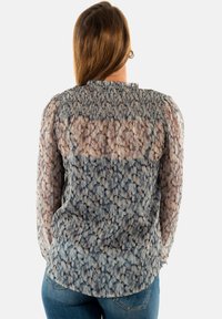 Blusa transparente con un canesú fruncido, que presenta un patrón floral en azul y beige, mangas largas abullonadas y un dobladillo curvado.