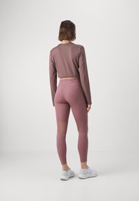 Femme debout de dos, portant un crop top mauve à manches longues, des leggings assortis avec des panneaux en maille, et des chaussures de sport blanches sur fond uni.