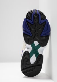 Vitt och svart atletisk sko sula med texturerat gummi, lila och gröna accenter, med detaljer i Torsion-teknologi och ett unikt mönster.