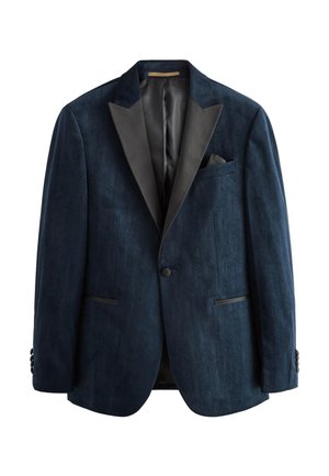 Mężczyzna w ciemnoniebieskiej aksamitnej marynarce smokingowej, czarnych spodniach garniturowych, białej koszuli, czarnej muszce, poprawiający rękawy, stojący na jednolitym szarym tle.