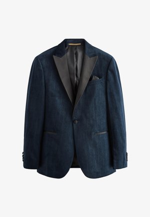 Mann in dunkelblauem Samtsmoking, schwarze Anzughose, weißes Hemd, schwarze Fliege, richtet Ärmel, steht vor einfarbigem grauem Hintergrund.