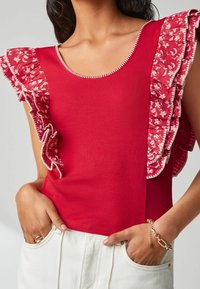 Débardeur rouge à côtes avec des volants à motifs floraux sur les épaules, des accents de couture blanche et un col rond.