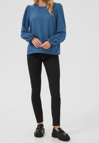 Blaue Bluse mit langen Ärmeln und gerafften Bündchen, kombiniert mit schwarzen, taillierten Hosen und klobigen schwarzen Slippern. Glatte Stoffoberfläche, lässige Passform.