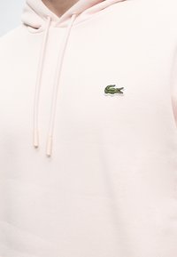 Sweat à capuche rose clair avec cordons et petit logo crocodile vert brodé sur la poitrine gauche.
