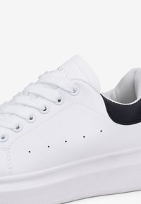 Sneaker bianco realizzato in pelle liscia, con un accento sul tallone in tessuto navy testurizzato, sei asole, lacci bianchi e piccoli fori di ventilazione.