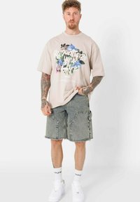 Lichtbeige oversized t-shirt met bloemenprint en tekst, gecombineerd met grijze denim shorts met een rafelrand en zijzakken. Witte sneakers.