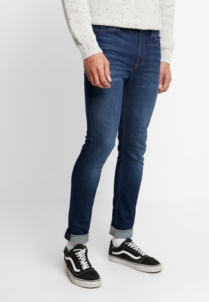 Mand iført mørkeblå slim-fit jeans med opkørte manchetter, hvide sokker, sort-hvide sneakers og en hvid strikket sweater, stående på en ensfarvet baggrund.