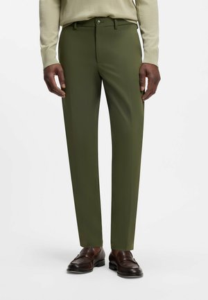 Mann trägt olivgrüne maßgeschneiderte Hose, beigen Pullover und dunkelbraune Lederloafers, steht vor weißem Hintergrund.