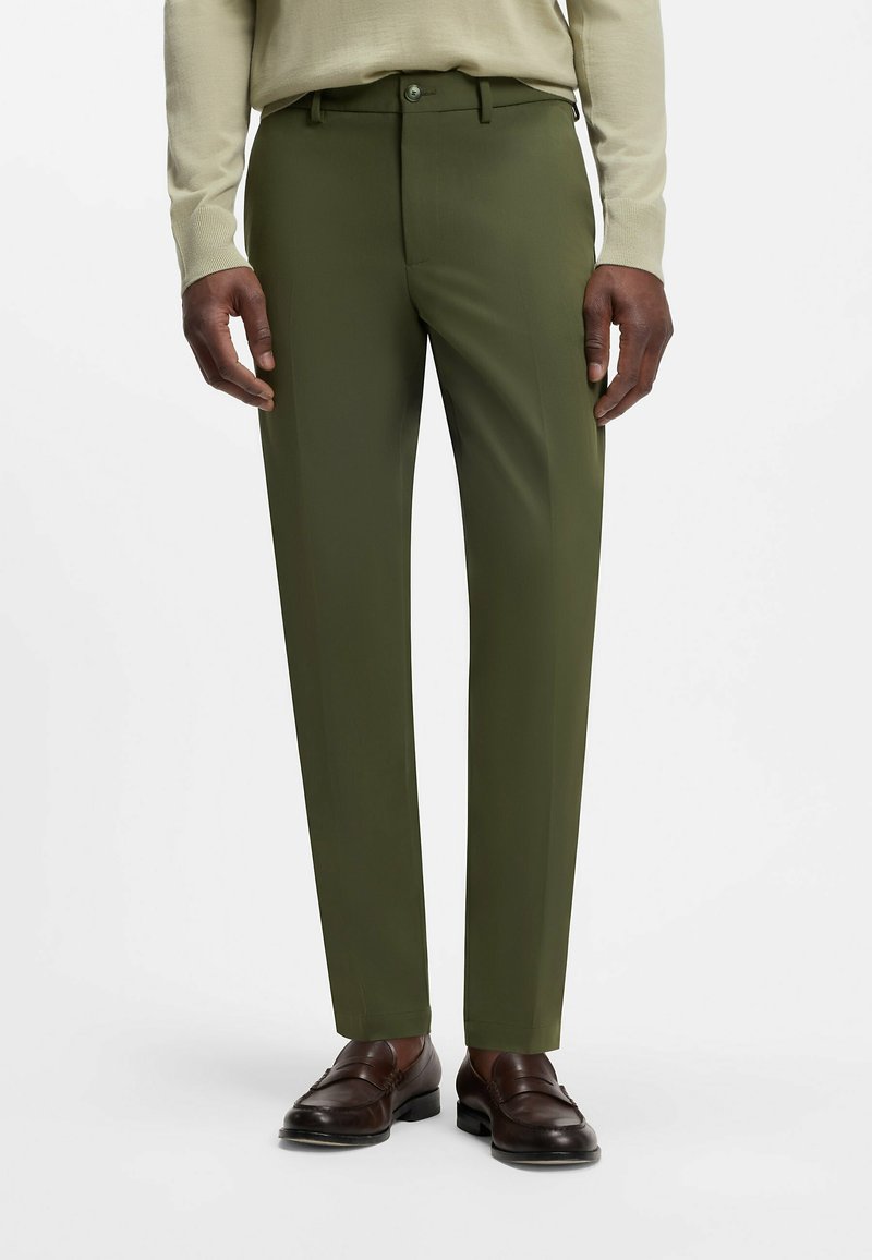 Homme portant un pantalon vert olive sur mesure, un pull beige et des mocassins en cuir marron foncé, debout sur fond blanc.
