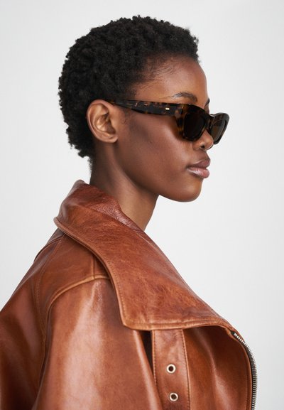 Tortoiseshell sunglasses with a pointed cat-eye shape, worn with a brown leather jacket featuring a wide collar and shiny texture.
