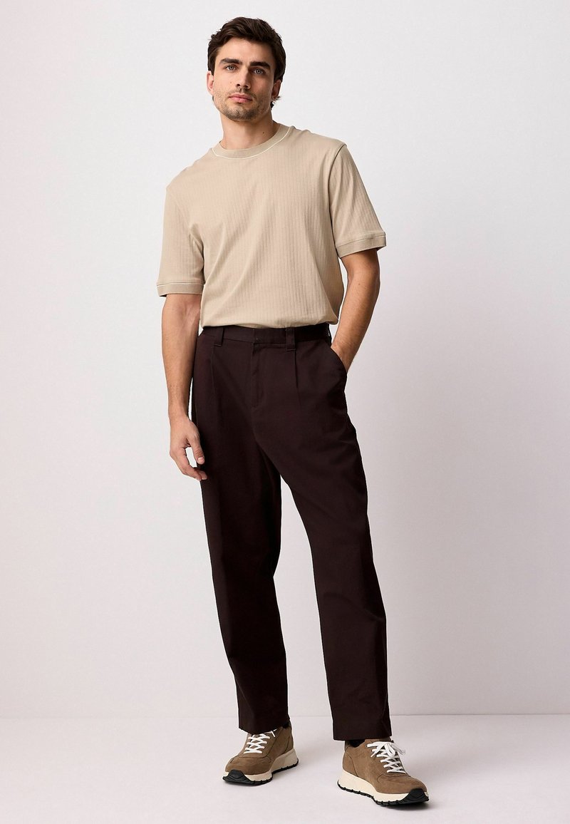Beige ribbet t-shirt med kortærmer, parret med mørkebrune bukser. Modellen har tan sko med hvide snørebånd på, og står foran en neutral baggrund.