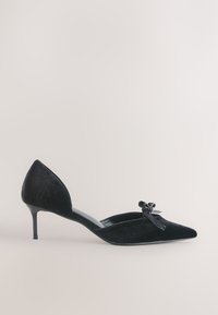 Chaussure en daim noire à bout pointu avec petit talon kitten heel, côtés ouverts et nœud décoratif sur le dessus, sur un fond clair uni.