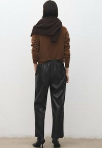 Brown knitted sweater draped over shoulders, paired with black leather trousers. Sleek texture, relaxed fit, and ankle-high black heels.