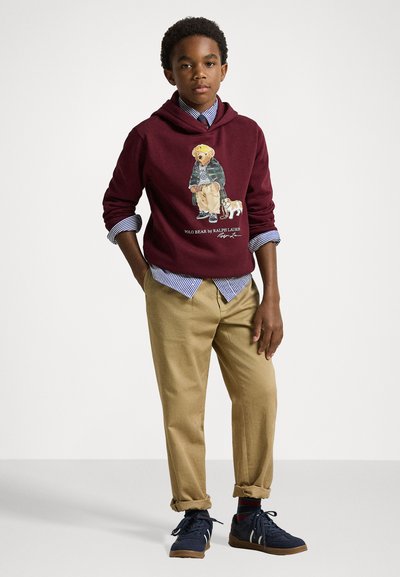 Bordo kapuca z dolgim rukavom s grafiko medveda, položen čez modro-belo črtasto srajco, v kombinaciji s bež hlačami in temnodβολ nošnimi čevlji. Gladka tekstura.