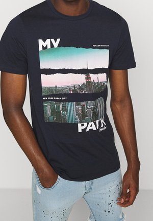 Homme portant un t-shirt noir avec une bande déchirée du skyline de New York et le texte "MY PATH" ainsi qu’un jean bleu clair avec des éclaboussures de peinture noire.