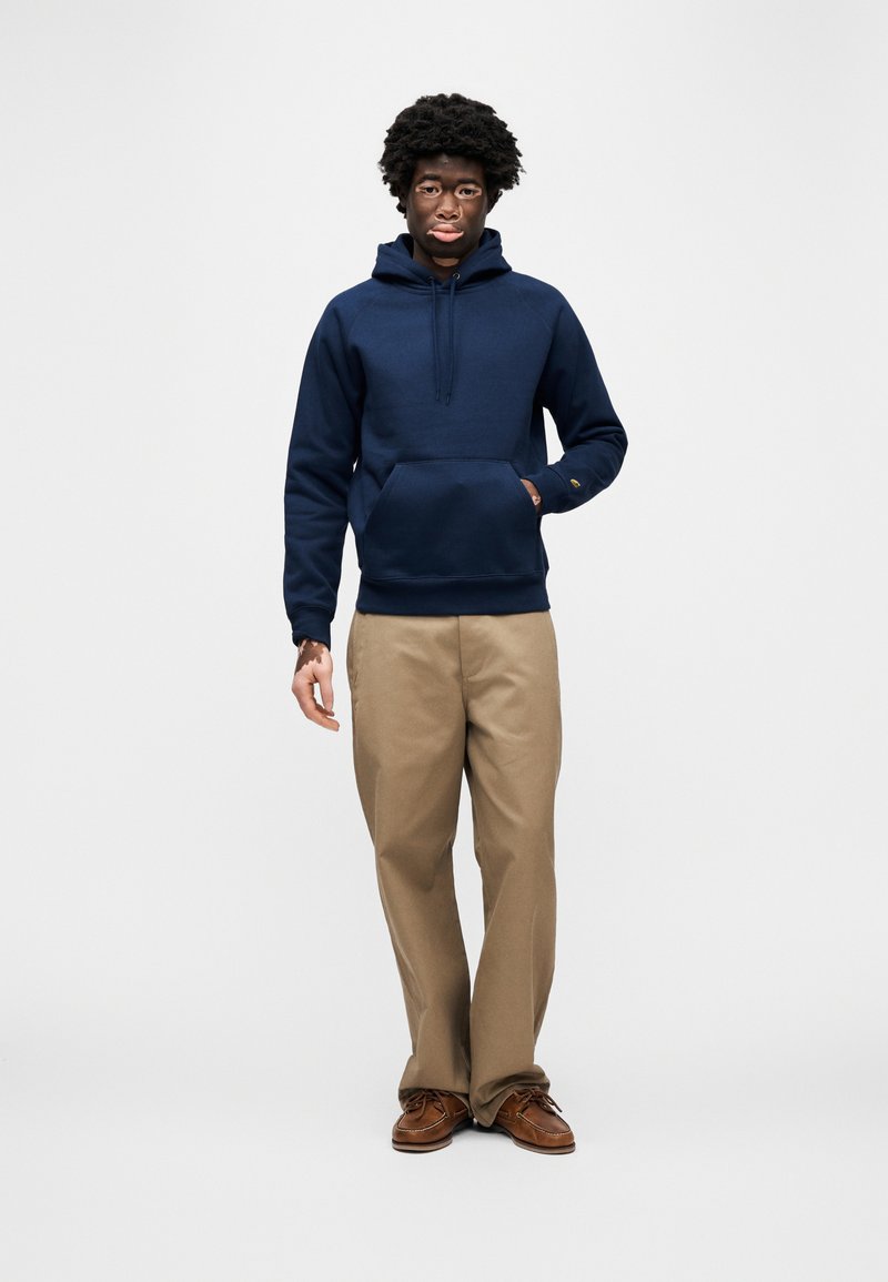 Sweat à capuche bleu marine avec poche kangourou et cordon de serrage, associé à un pantalon kaki et des chaussures marron, présenté sur un fond uni.