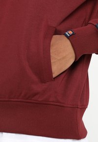 Hand in de zak van een bordeauxrode sweatshirt met een bijpassende label waarop het Ellesse-logo staat, ribbelzoom en gedeeltelijk zicht op een wit onderhemd.