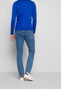 Chemise bleue à manches longues, jean slim en denim, et baskets blanches avec des accents bordeaux. Le tissu semble lisse avec un design classique de poche arrière.