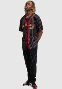 Svart pinstripet t-skjorte med korte ermer og røde detaljer samt trykk av 'KANI 1989', kombinert med svarte bukser med skrå hvite sømmer og røde sneakers.