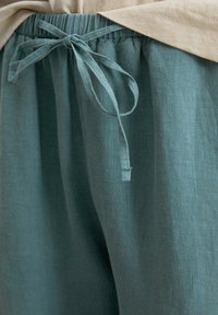 Teal linnen broek heeft een elastische taille met strik-touwtjes. De stof heeft een zachte, gestructureerde afwerking, met plooien en val.