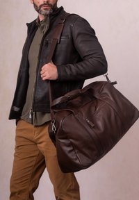Brown leather duffle bag with a textured finish, featuring a detachable shoulder strap and top handles, carried by a person in casual attire.