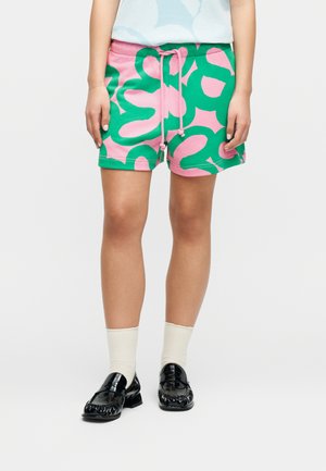 Osoba oblečená v ružovo-zelených šortkách s abstraktným vzorom, bielych ponožkách a čiernych lesklých topánkach, stojaca pred jednofarebným pozadím.