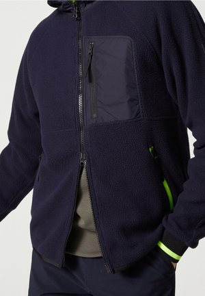 Homme portant une veste polaire bleu marine avec poche poitrine zippée et deux poches latérales, dont une avec un détail de fermeture éclair vert néon, par-dessus une chemise olive.