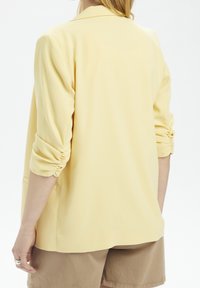 Light yellow blazer with a relaxed fit, ruched sleeves, and a smooth texture. Features a classic collar and a straight-cut back.