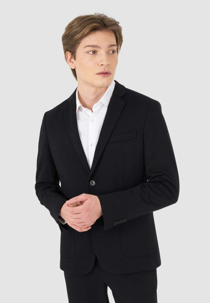 Giacca nera con rever a taglio e una tasca sul petto; abbinata a una camicia bianca. Il tessuto sembra liscio e ben tagliato per un look formale.