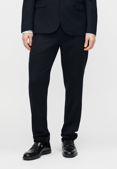 Άνδρας που φοράει μαύρο παντελόνι κοστουμιού, μαύρα δερμάτινα παπούτσια και ταιριαστό μαύρο σακάκι, με τα χέρια χαλαρά στα πλάγια.