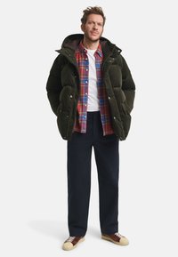 Mann trägt eine dunkelgrüne Fleecejacke, ein rotkariertes Hemd, ein weißes T-Shirt, dunkle Hosen und braune Sneakers und steht mit den Händen in den Taschen.