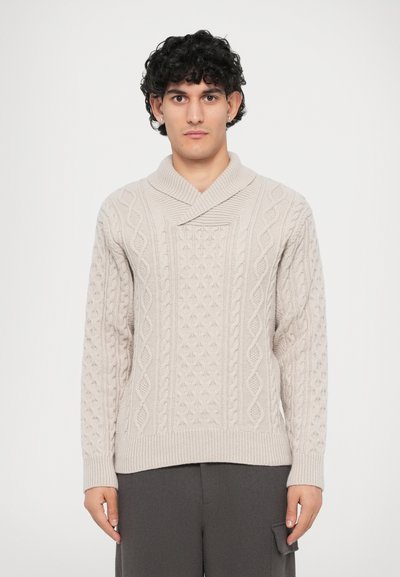 Pull en maille câble crème avec col châle, présentant des motifs texturés et des poignets côtelés, porté sur un pantalon gris foncé.