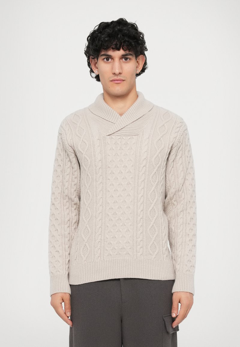 Krém színű, cable-knit pulóver sálgallérral, textúrált mintákkal és bordás mandzsettákkal, sötétszürke nadrág fölé öltözve.