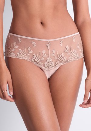 Femme portant des sous-vêtements en dentelle rose pâle transparente avec broderie florale, debout devant un fond uni neutre.