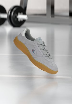 Adidaș gri cu talpă de gumă și logo, plutind deasupra unei podele albe reflectorizante, cu greutăți de sală de sport neclare în fundal.