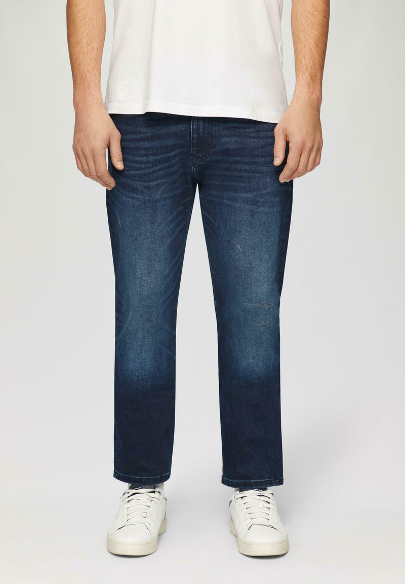 Jeans en denim bleu foncé avec une coupe droite, présentant des détails de décoloration subtile et de moustaches, associés à des baskets blanches.